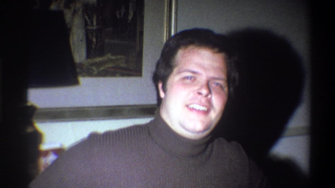 Smiling young man in a dark turtleneck facing the camera with a large shadow on the wall.