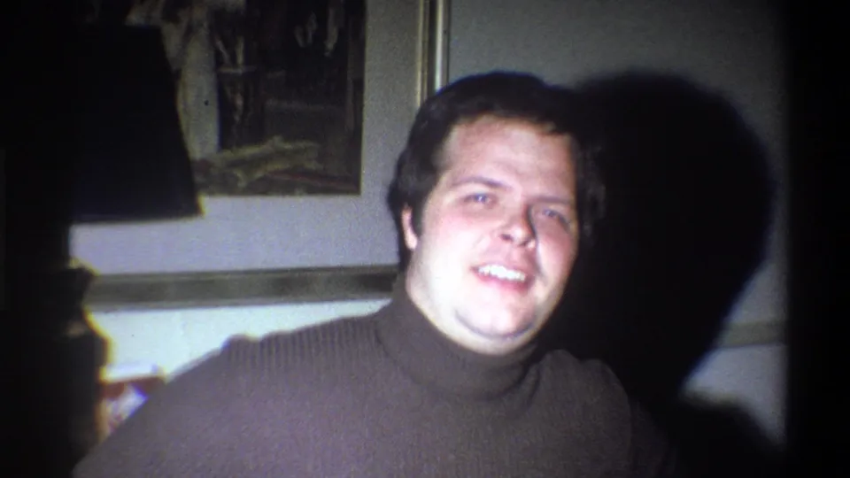 A young man in 1973 Troy, New York home footage smiles toward the camera, with a strong shadow cast behind him indoors.