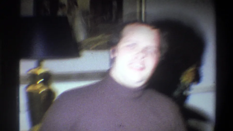 A blurred 1973 Troy, New York frame shows the man moving in a dim private interior near a lamp and wall decor.