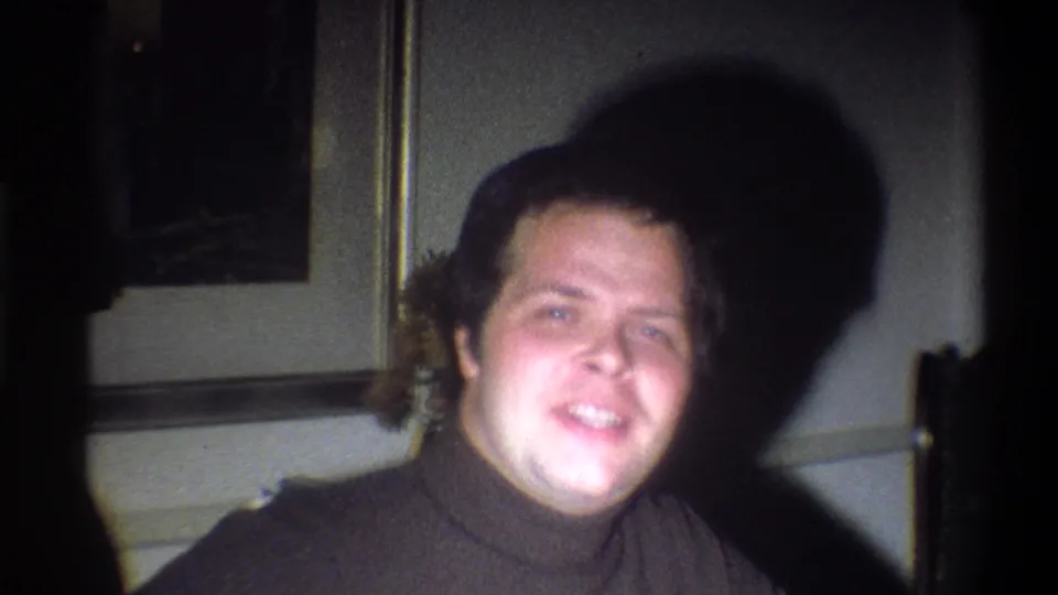The 1973 Troy, New York home-movie frame shows the man facing the camera in a dark turtleneck, with framed art and shadow behind him.