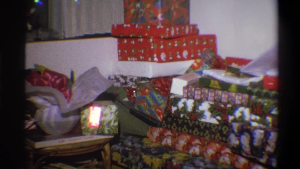 1973 Troy, New York home-movie frame showing densely stacked Christmas gifts and a bright light on one foreground package.