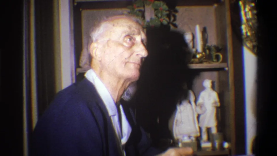 An elderly man, likely the grandfather noted in metadata, appears in a 1973 Troy, New York home interior near shelves and small figurines.
