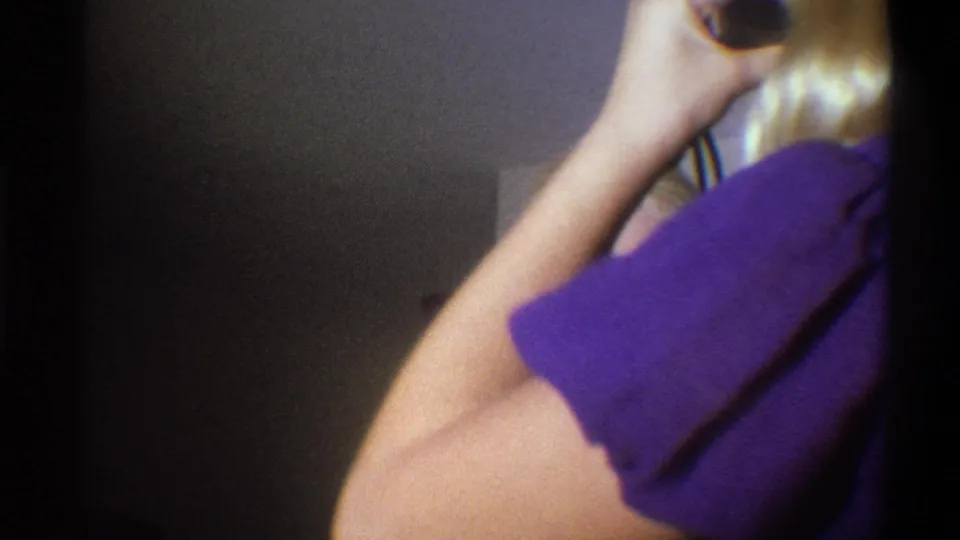 A blonde woman in a purple top raises a handheld camera near her face in soft-focus 1973 Troy home-movie footage.