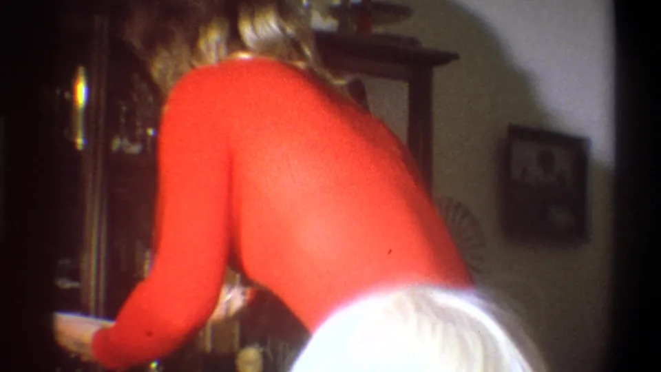 A blonde woman in a red sweater bends forward in a private interior, with furniture and wall decor visible behind her.