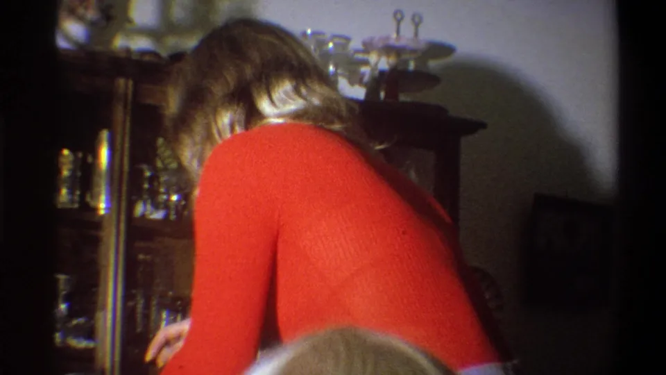 In a 1973 Troy, New York home interior, a blond woman in a red long-sleeved shirt bends near a wooden china cabinet.