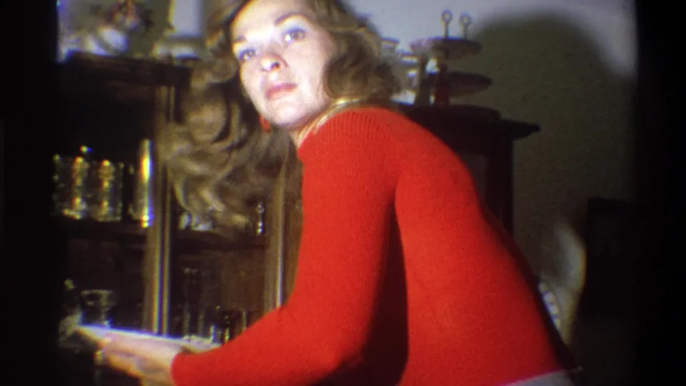 The blond woman turns toward the camera in the 1973 Troy interior while holding a paper plate near the china cabinet.