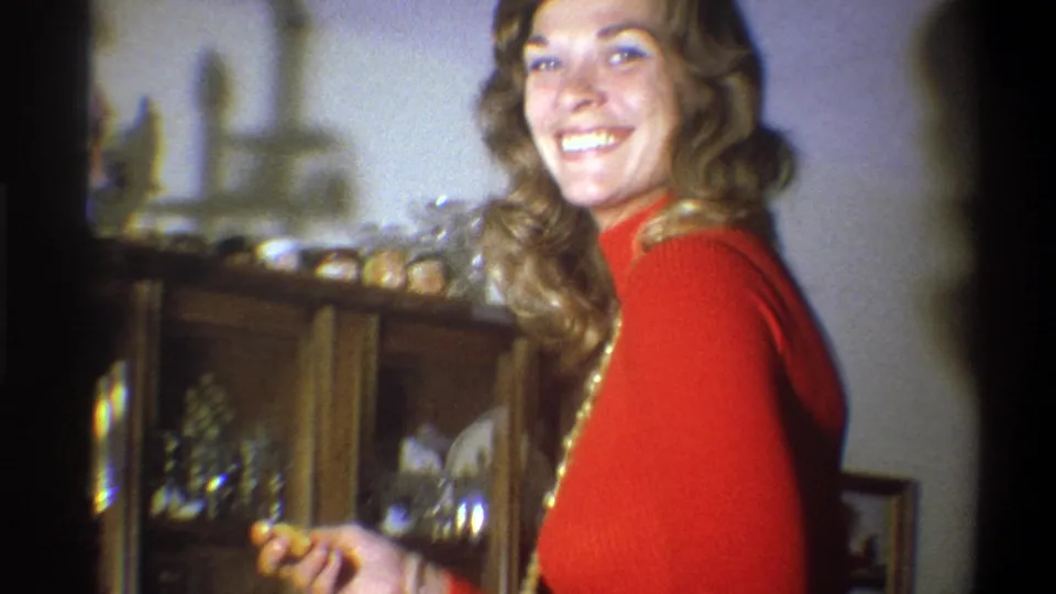 A smiling blond woman in a red shirt looks toward the camera inside the 1973 Troy home, holding a paper plate by the cabinet.
