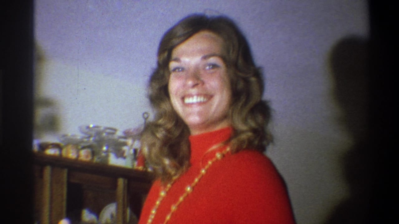 In a 1973 Troy, New York home interior, a woman in a red turtleneck smiles near a wooden shelf holding glassware.