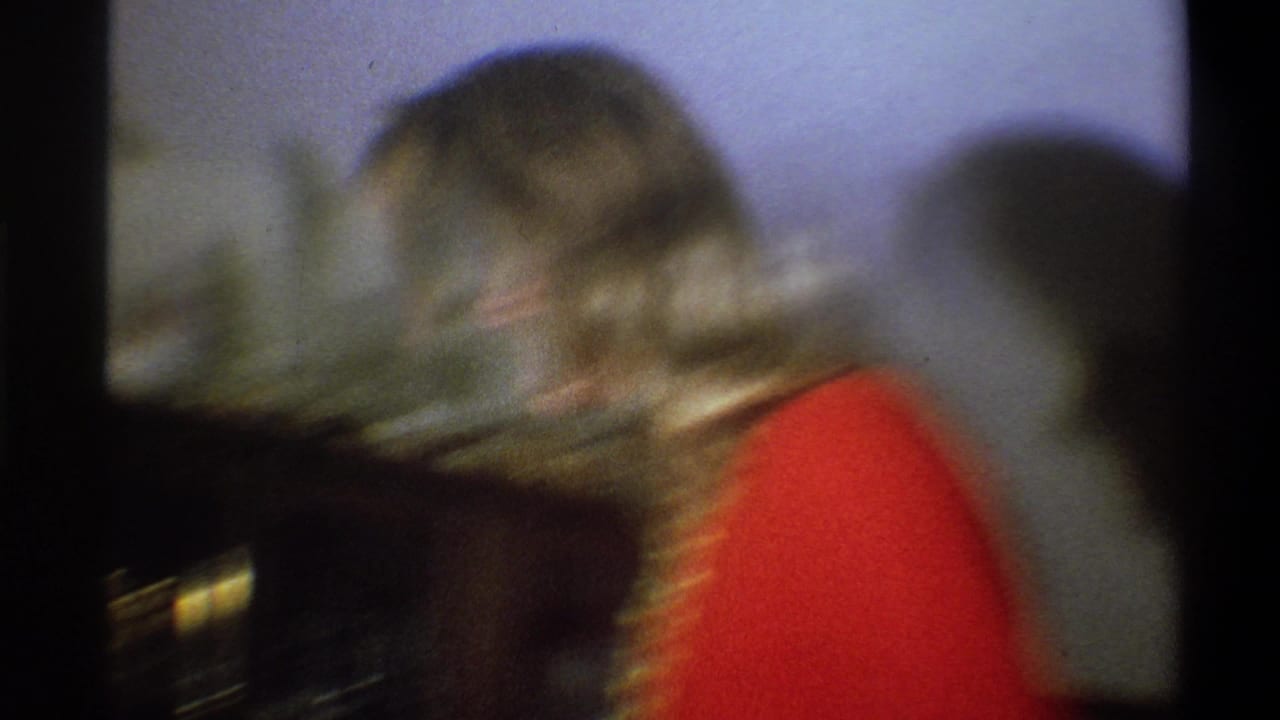 Soft, blurred 8mm frame of the woman in red moving near the shelf inside the Troy home interior.