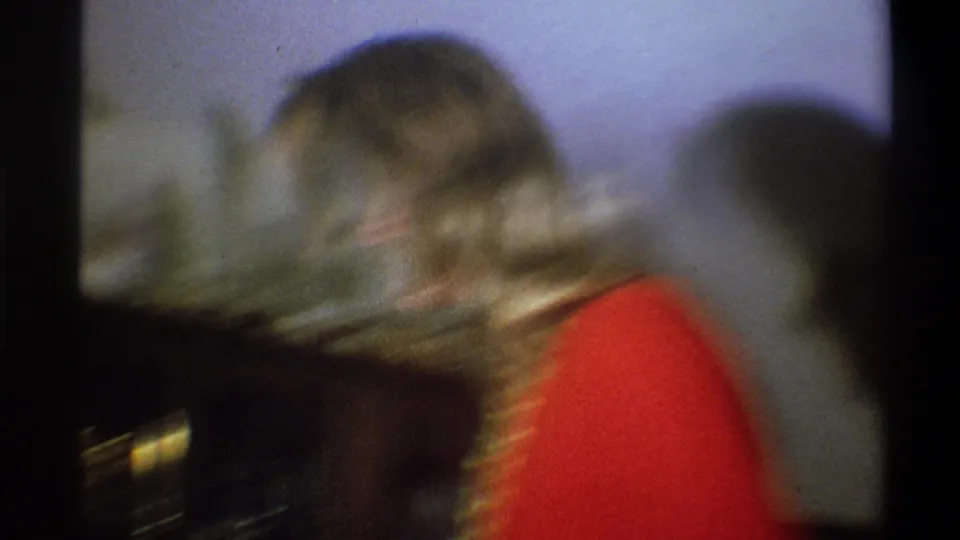 Soft, blurred 8mm frame of the woman in red moving near the shelf inside the Troy home interior.