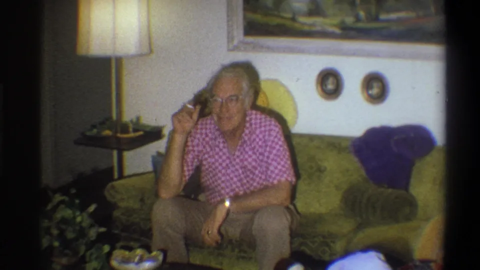 Older man in a pink checkered shirt sits on a green sofa in a 1973 Troy home, holding a cigarette near a lamp and wall art.