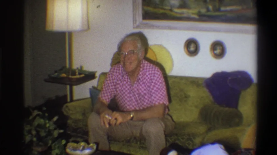 Older man seated on a green sofa in the Troy home interior, with a lamp, framed painting, wall plates, and cushions nearby.