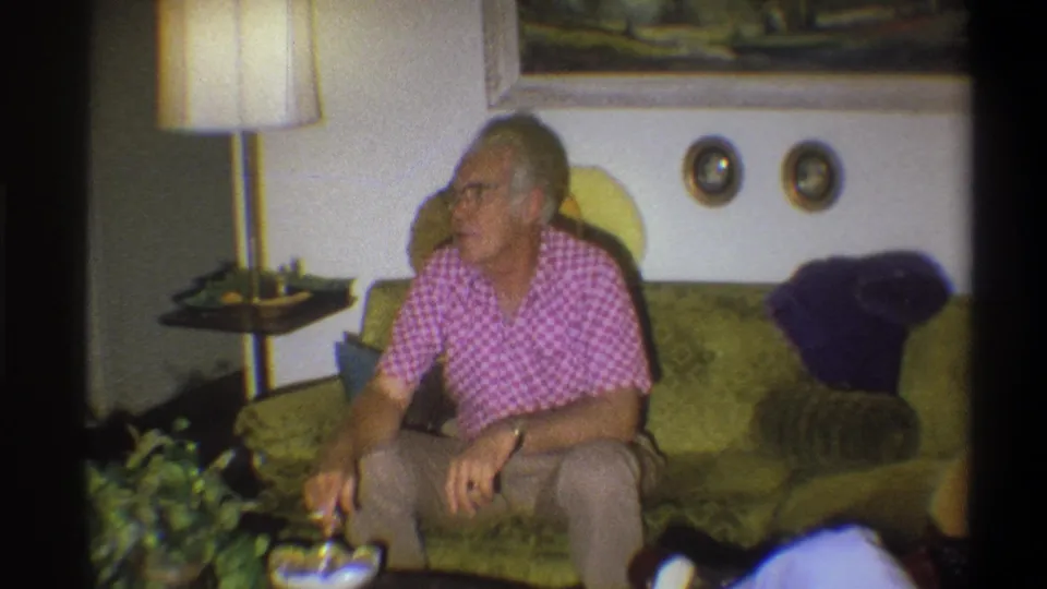 The elderly man remains seated on the green couch, turned toward the side of the room in the Troy, New York living-room scene.