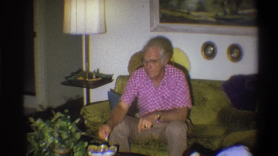 A 1973 home-movie frame shows the man leaning forward on the green couch, with a houseplant, lamp, and framed decor around him.