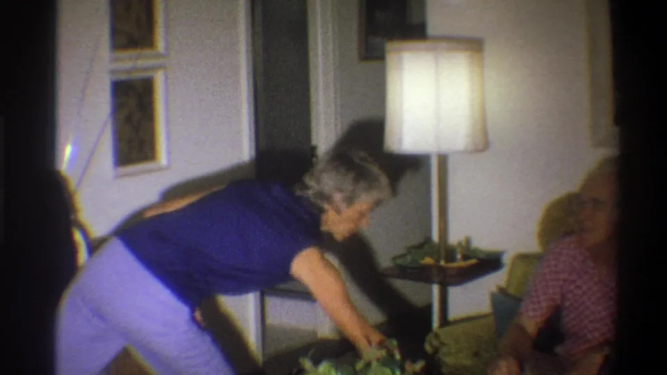 A woman in a blue top reaches into the living room as the elderly man sits partly at the edge of the frame.