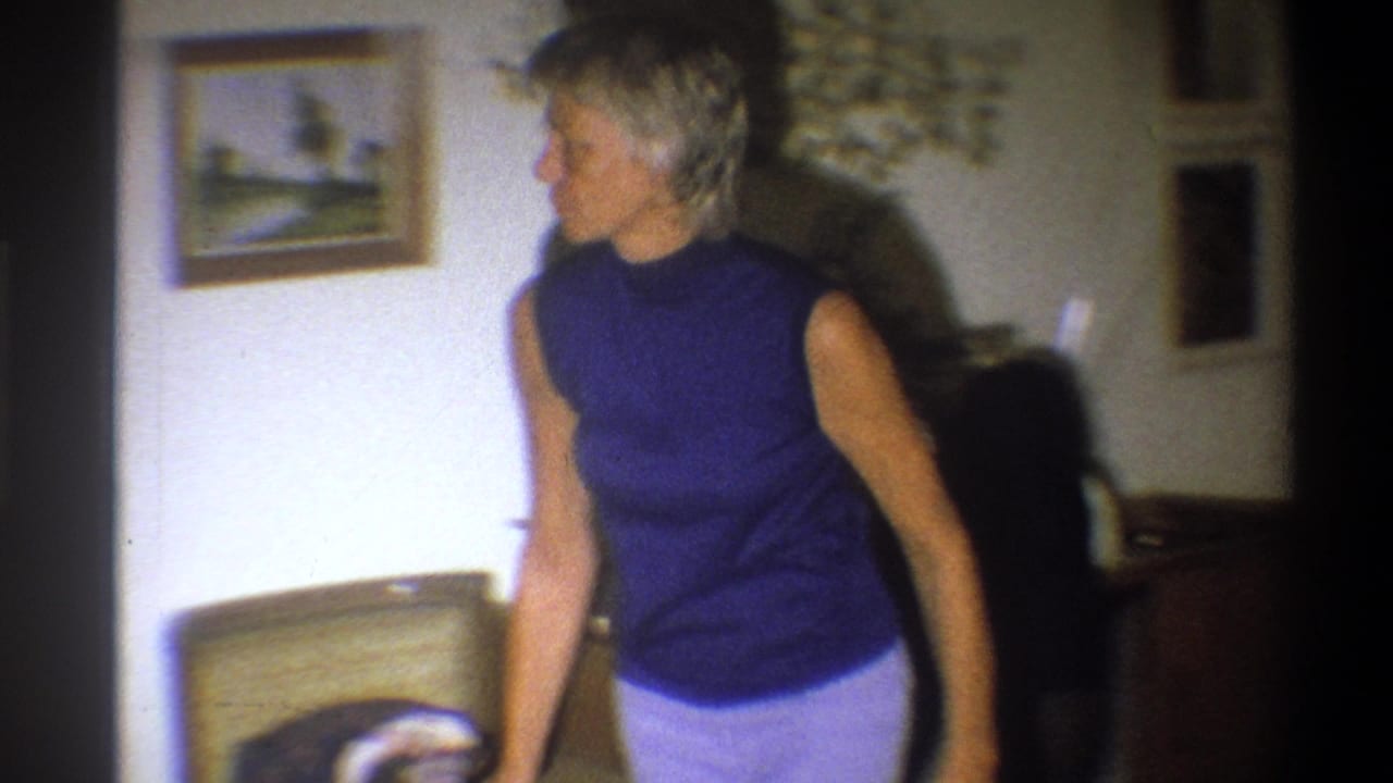 Woman in a blue sleeveless top stands beside a chair in a living room.