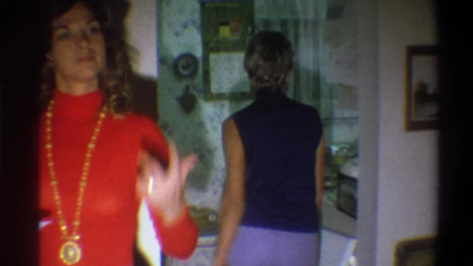 Inside the Troy home, a woman in a red turtleneck and long necklace stands near the camera while another woman enters a floral-wallpapered kitchen.