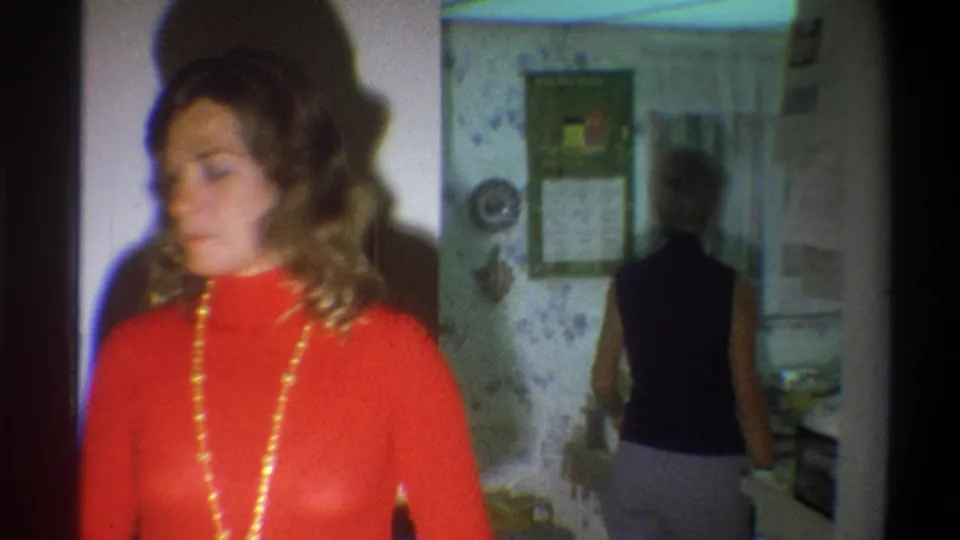 A woman in a red turtleneck and necklace stands close to the camera as another woman moves away through the 1973 Troy kitchen interior.