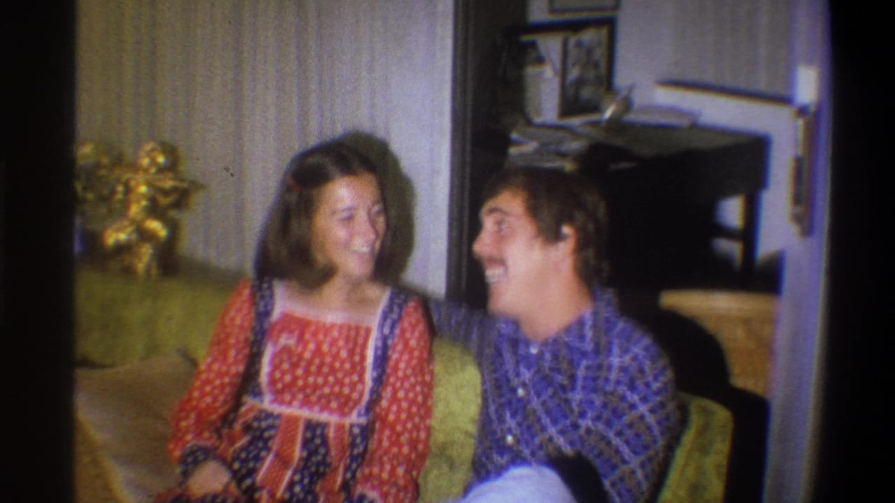 The couple laughs together on a green couch, with 1970s wallpaper and household decor visible in the Troy interior.