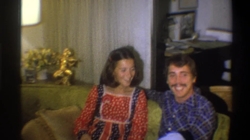 A woman in a red patterned dress looks down as a man in a blue plaid shirt faces the camera in the 1973 Troy living room.
