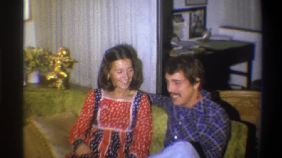 The young couple smiles at each other on a green couch, framed by 1970s living room decor in Troy, New York.