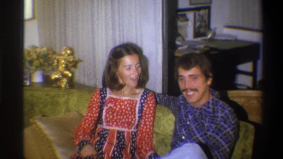 In 1973 Troy, New York home-movie footage, a young couple sit close together on a green couch in a private living room.