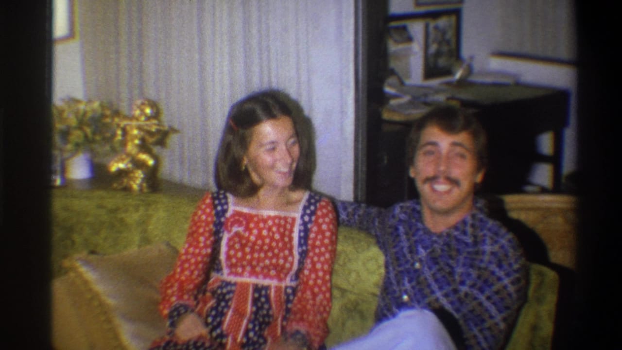 The couple smile in a 1973 Troy, New York living room, seated close together on a green couch with household decor visible.