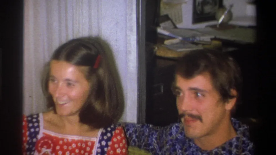 A closer 8mm frame shows the couple laughing, with the woman turned aside and the man glancing toward her in a Troy living room.