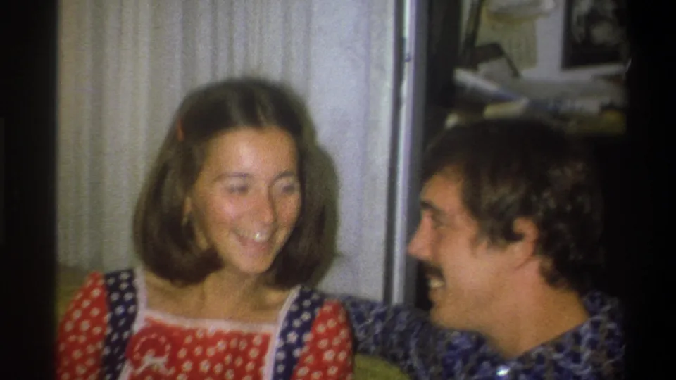 In the 1973 home-movie interior, the seated pair laugh together as the man looks toward the woman beside him.