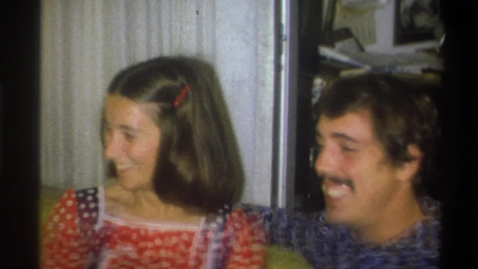 A young couple in 1973 Troy, New York footage laugh face to face, framed by curtains and the soft grain of 8mm film.