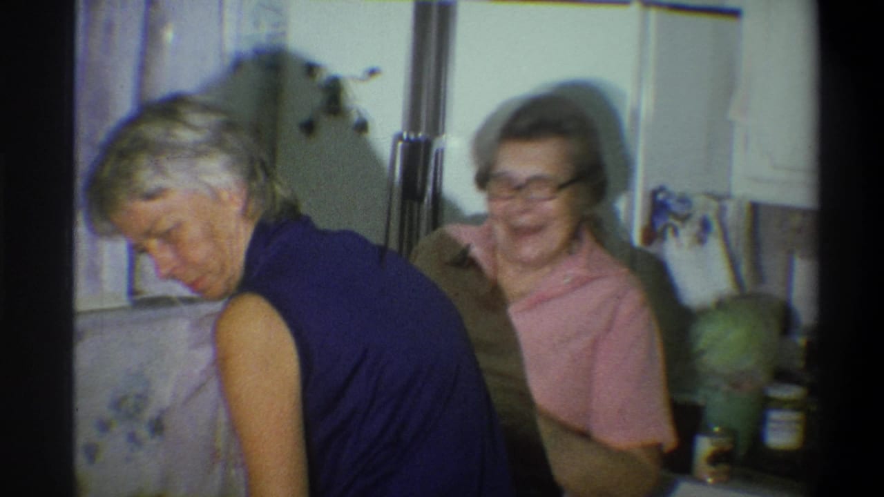 In the 1973 Troy kitchen scene, a woman in a blue sleeveless top bends forward as a woman in glasses smiles behind her.