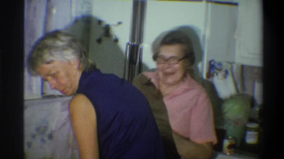 In the 1973 Troy kitchen scene, a woman in a blue sleeveless top bends forward as a woman in glasses smiles behind her.