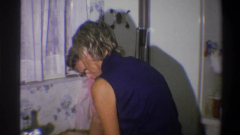 In a 1973 Troy, New York home kitchen, a woman in a dark sleeveless top bends toward the counter beside floral wall decor.