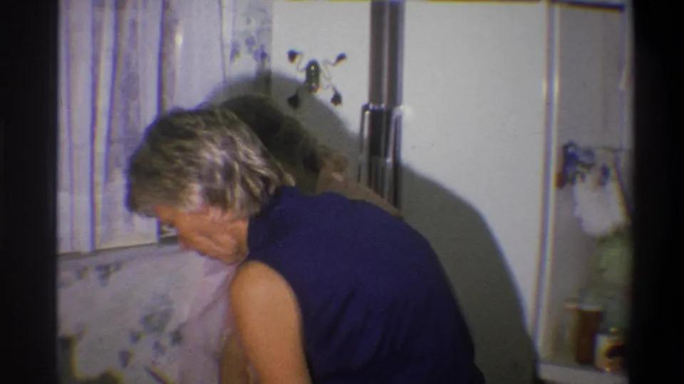 A dark-topped woman bends over the kitchen work area in 1973 Troy, New York home-movie footage.