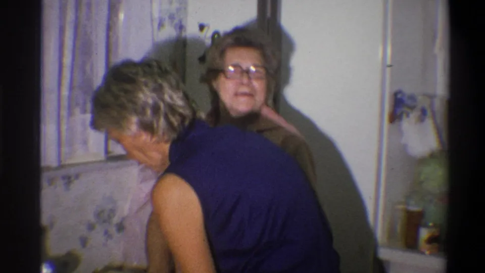 A woman in glasses and a pink top smiles toward the camera behind another woman in a 1973 Troy home kitchen.