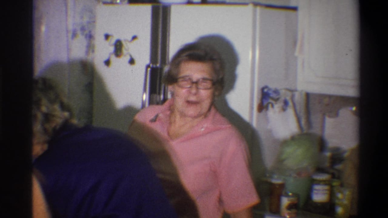 In a 1973 Troy, New York home-movie frame, an older woman in glasses and a pink shirt smiles beside a refrigerator with magnets.