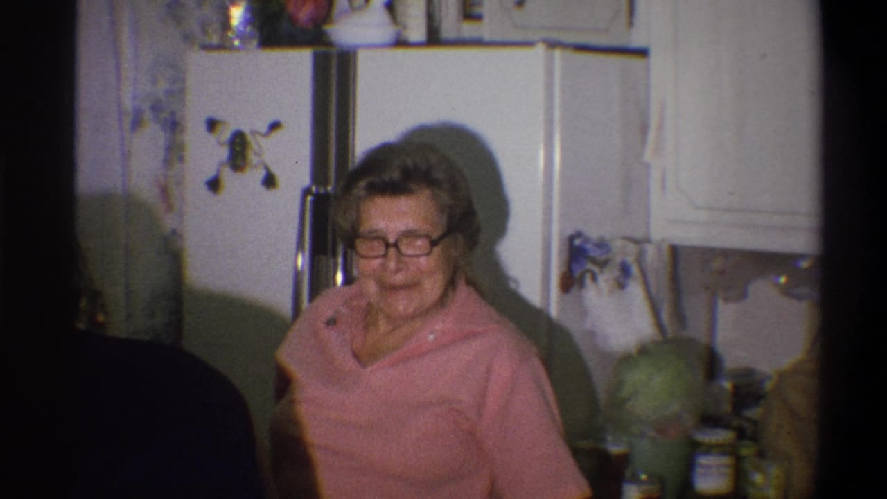 The older woman remains near the white refrigerator, smiling toward the camera in the Troy kitchen interior.