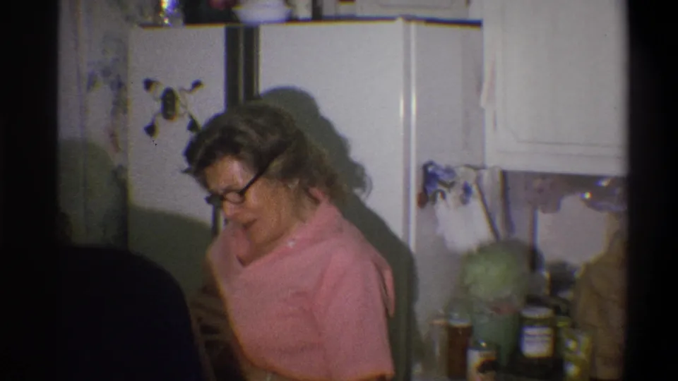 The woman turns downward beside the refrigerator, with jars and cans visible on the counter in the 1973 kitchen scene.