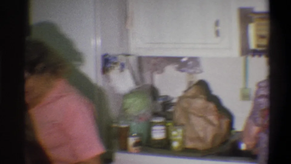 The woman moves out of frame in the Troy kitchen, leaving the counter, jars, cans, paper bags, and refrigerator area prominent.