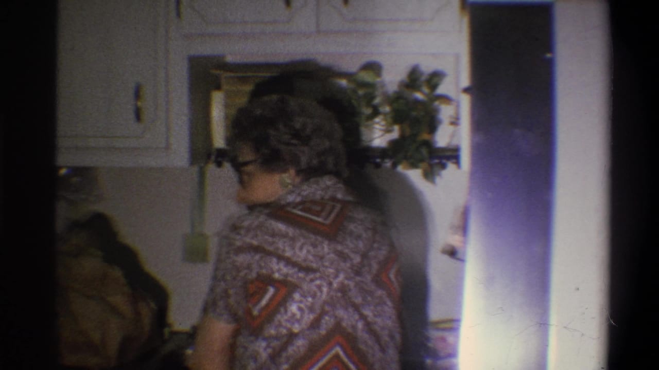 1973 Troy, New York home-movie frame showing an older woman in glasses near a kitchen sink, with white cabinets and a hanging plant visible.