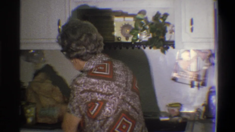 1973 Troy, New York home-movie frame showing the woman from behind at the kitchen sink beneath white cabinets and a wall shelf area.