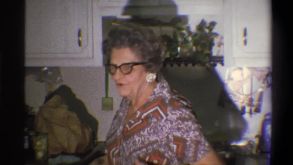 1973 Troy, New York frame of an older woman in glasses turning toward the camera in a private kitchen interior.