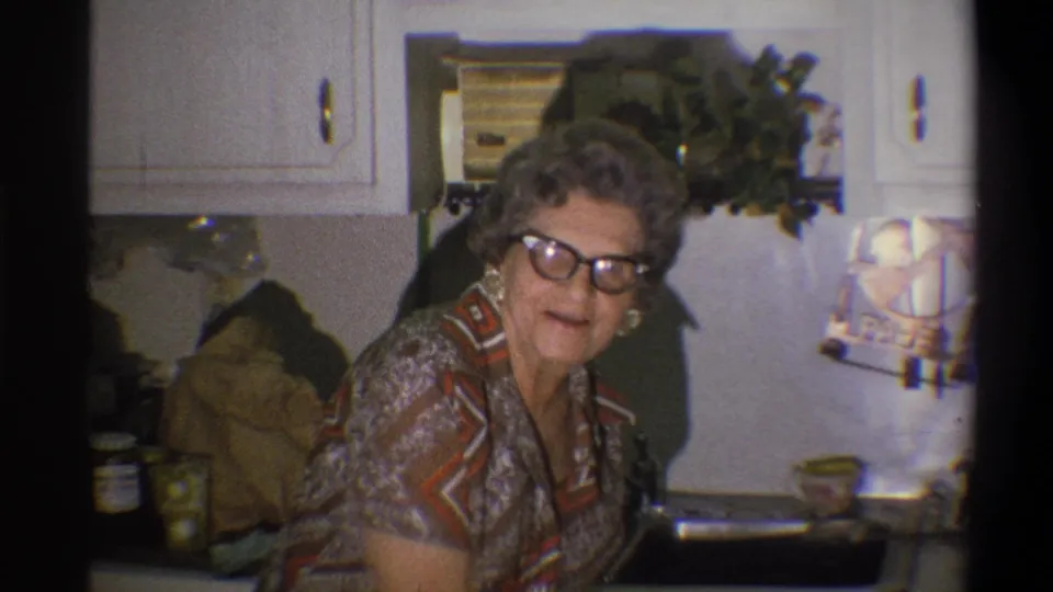 Older woman wearing glasses and earrings looks toward the camera in a 1973 Troy kitchen interior.
