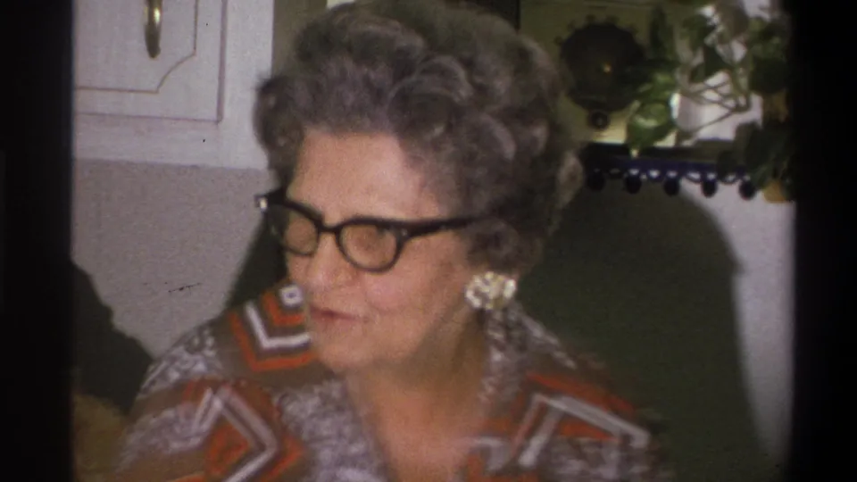 Close 8mm frame of an older woman with glasses, large earrings and a patterned blouse in a Troy kitchen.