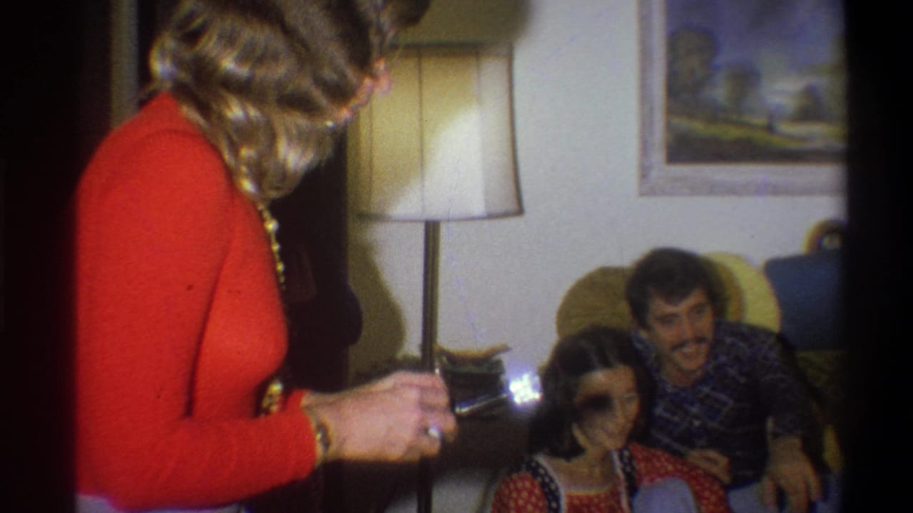 In a 1973 Troy, New York home-movie frame, a woman in an orange dress photographs a seated couple in a dim living room.