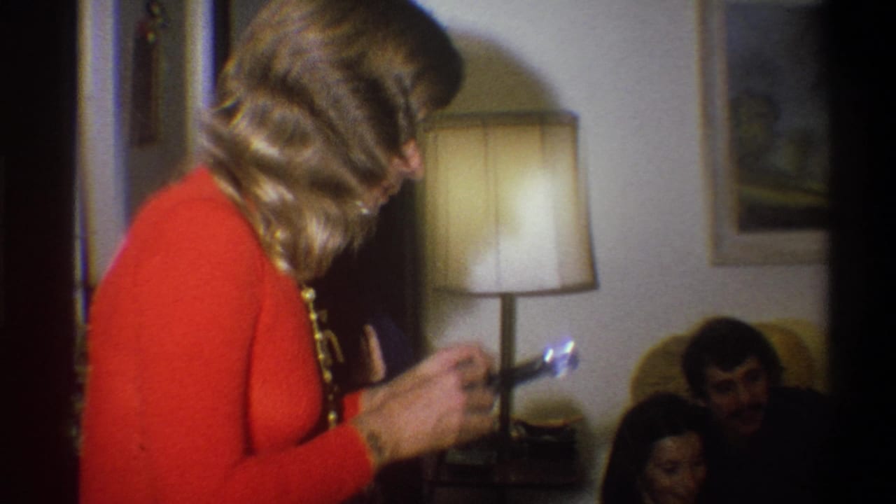 Woman in an orange dress aims a flash camera toward people seated near a lamp.