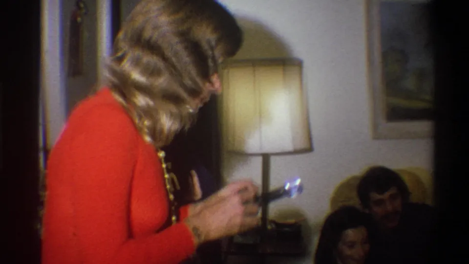 The woman in orange turns with a small flash camera toward seated people in a Troy living room, with a lamp and wall art behind them.