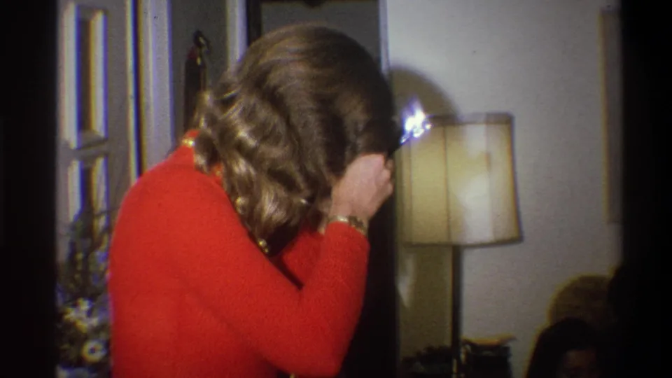 A 1973 home-movie frame shows the woman bringing the flash camera close to her face in the living room, partly obscuring seated people.
