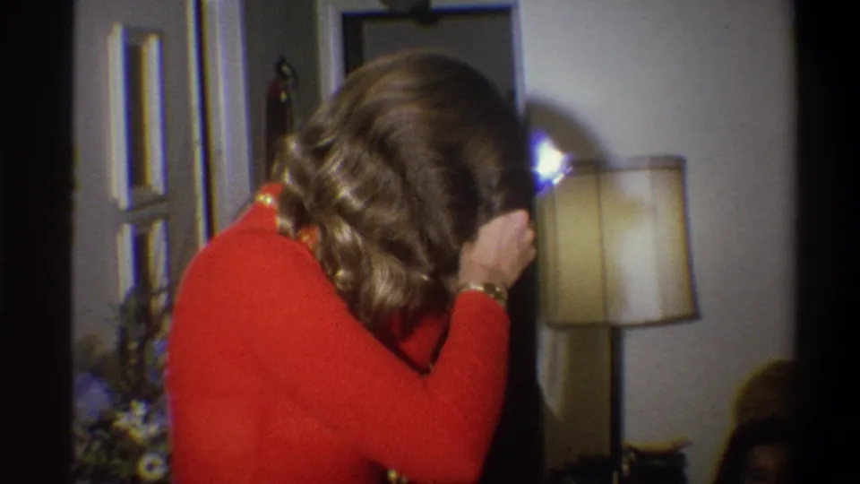 The woman holds the camera near her face as a bright flash reflection appears beside the lamp in the Troy living-room footage.