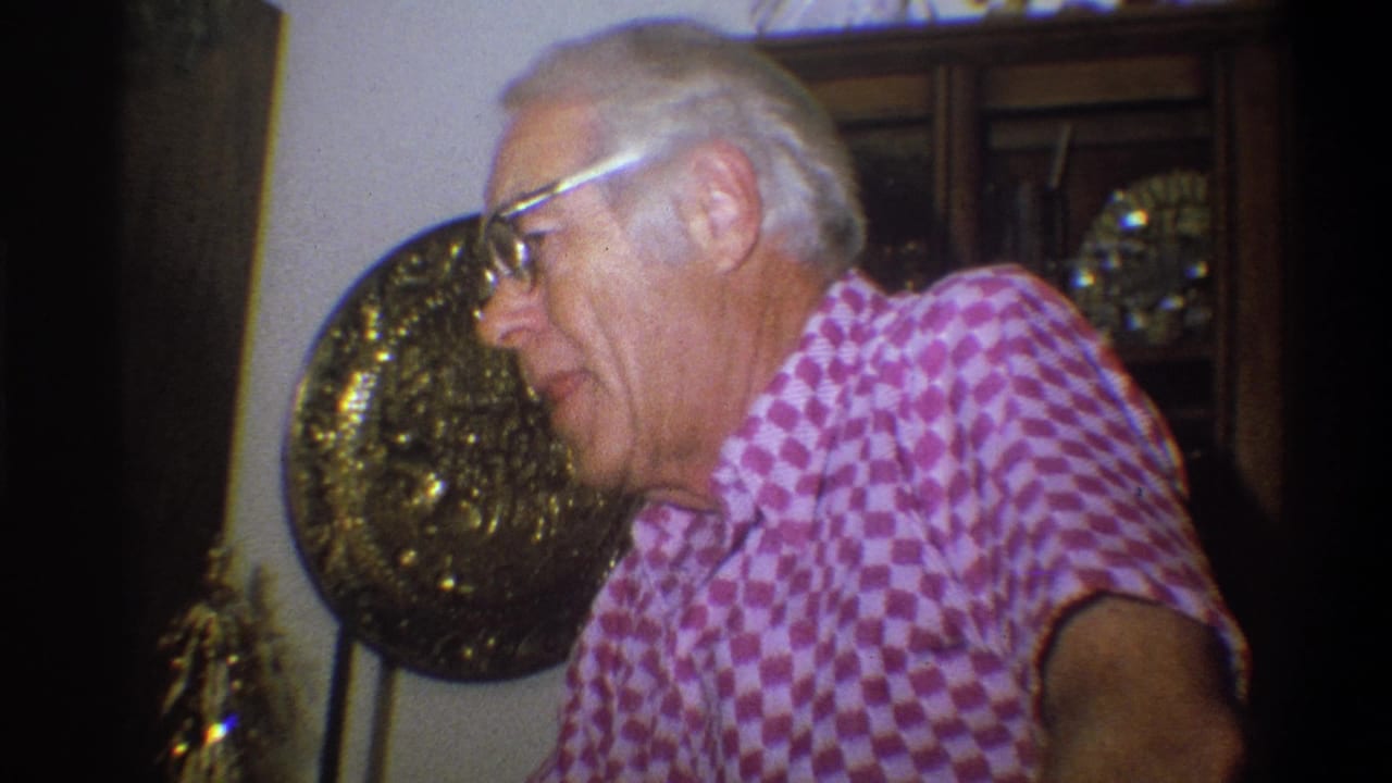 Older man in glasses leaning forward in a pink checkered shirt indoors.
